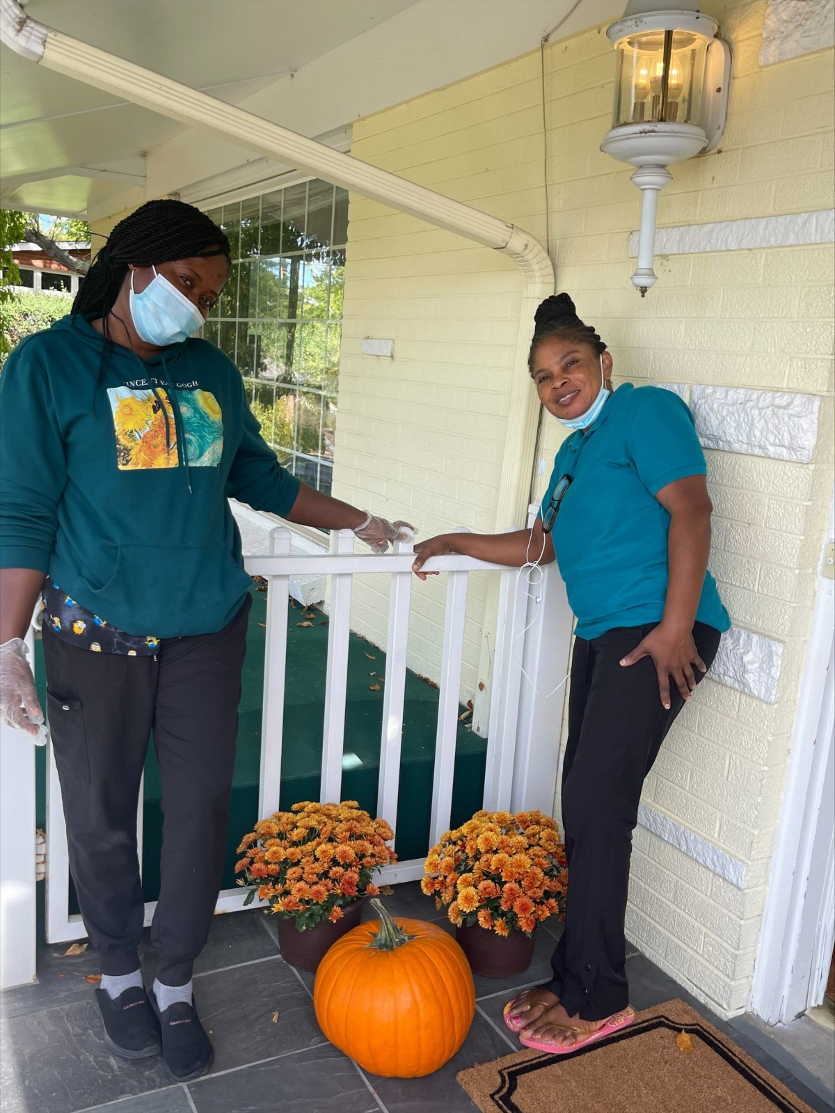 Two individuals making a delivery on a porch decorated with flowers and a pumpkin, both wearing masks and gloves.