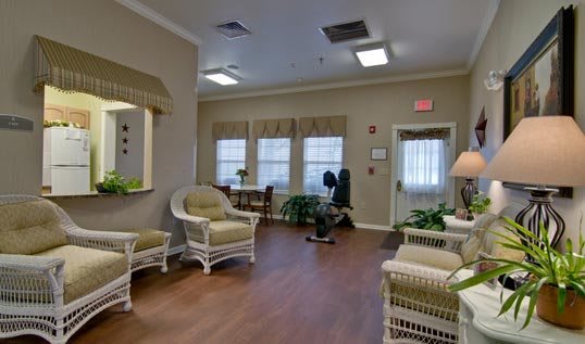 A cozy living room with wicker furniture and plants, featuring large windows that bring in natural light.