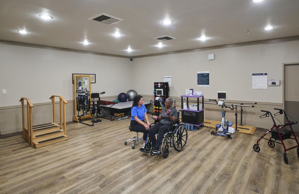 A therapist assists a patient in a wheelchair within a rehabilitation facility equipped with various exercise machines and therapy tools.