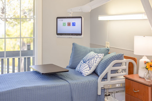 A modern hospital room featuring a patient bed with blue bedding, decorative pillows, and a wall-mounted monitor.