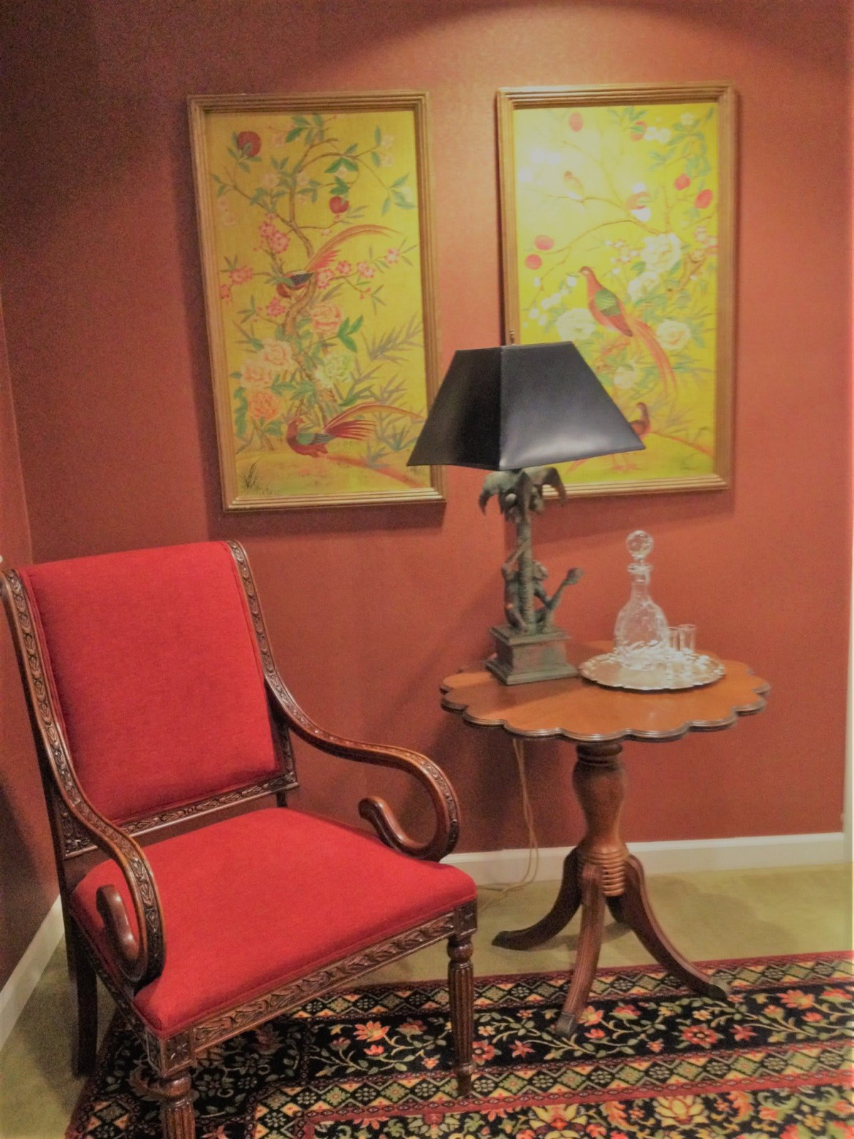 A cozy corner featuring a red upholstered chair, a small wooden table with an ornate lamp, and two decorative paintings on a warmly-colored wall.