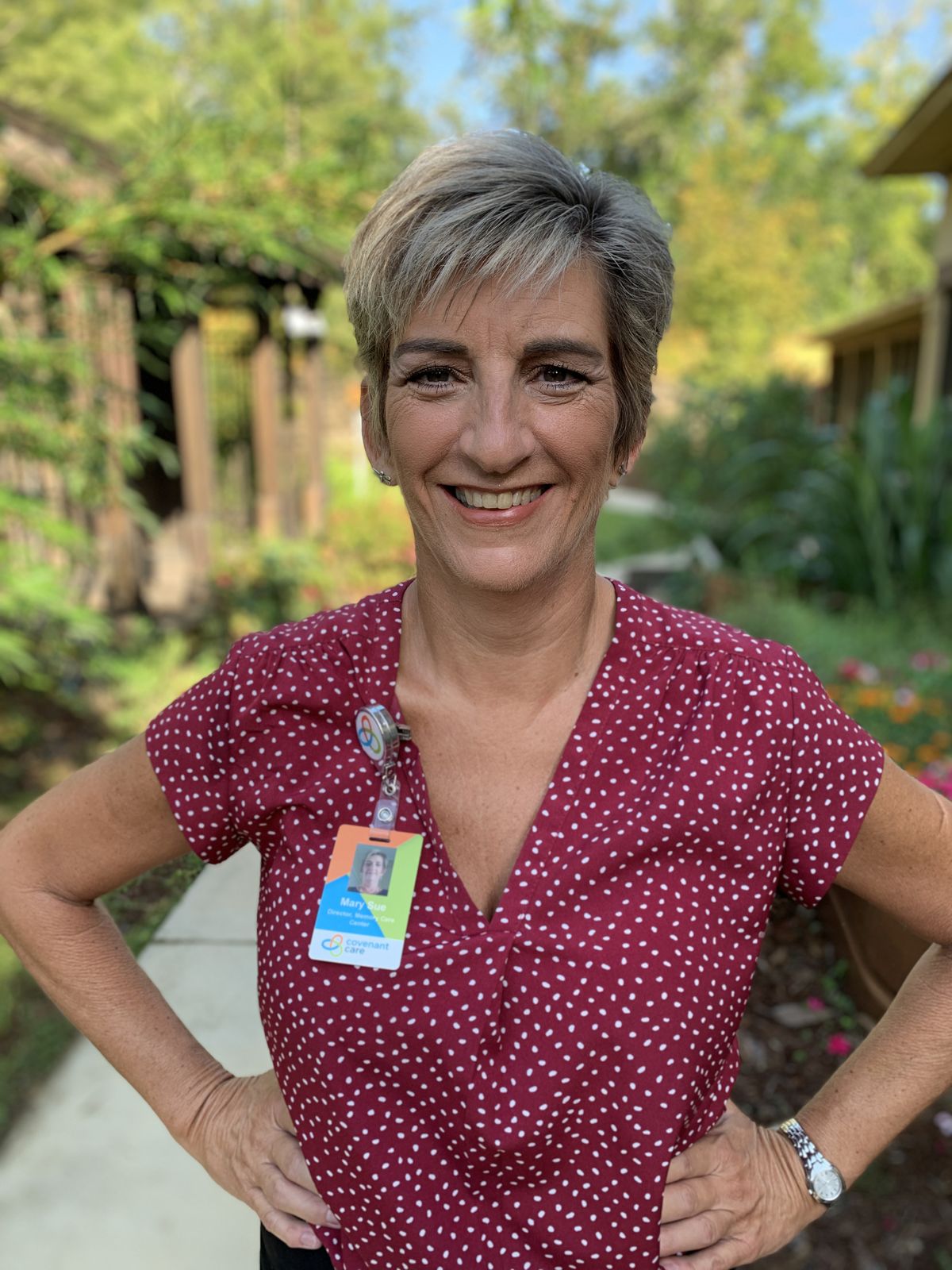 A woman with short hair, wearing a polka dot shirt and an ID badge, smiles while standing in a garden.