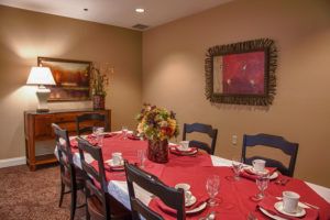 A warmly lit dining room with a table set for guests, featuring a red tablecloth and floral centerpiece.
