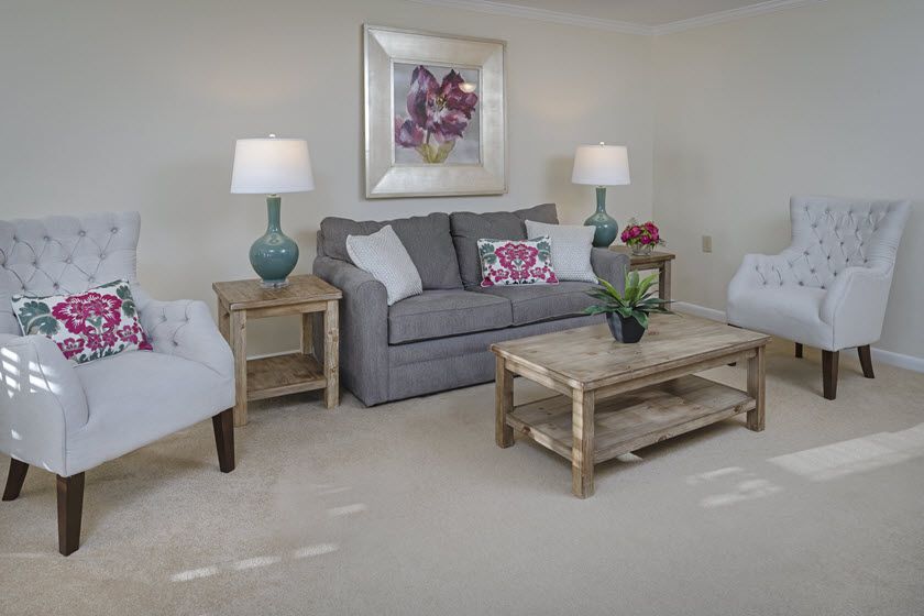 A cozy living room with a gray sofa, armchairs, and wooden furniture, decorated with lamps and a colorful painting.