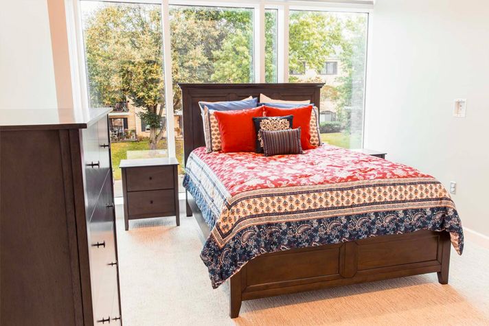 A brightly lit bedroom with a colorful bedspread and large windows.