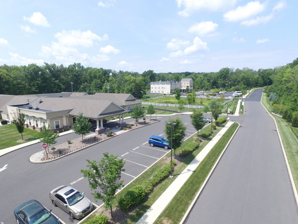 Aerial view of a parking lot with multiple cars and buildings surrounded by trees under a partly cloudy sky.