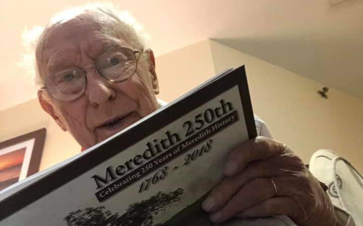 An elderly person wearing glasses is reading a booklet titled 'Meredith 250th'.