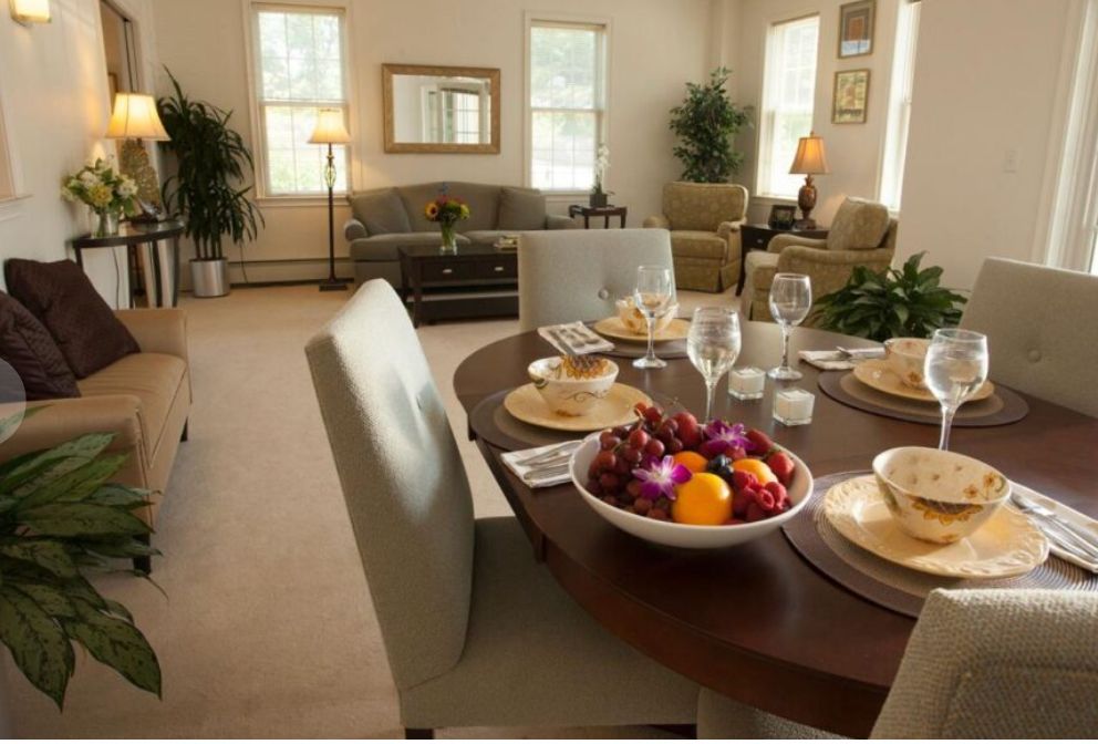 A modern living and dining area with a round table set with plates, glasses, and a fruit bowl, surrounded by comfortable chairs.