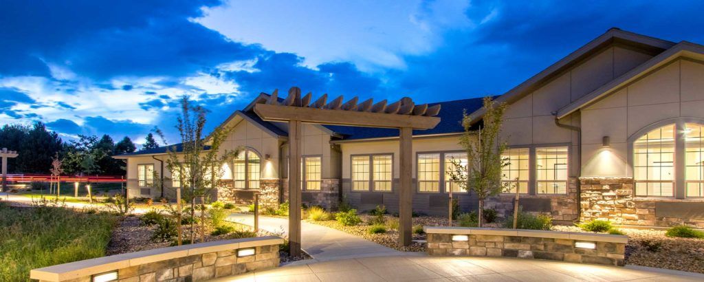 A well-lit house exterior with a pergola at night, surrounded by landscaped gardens.