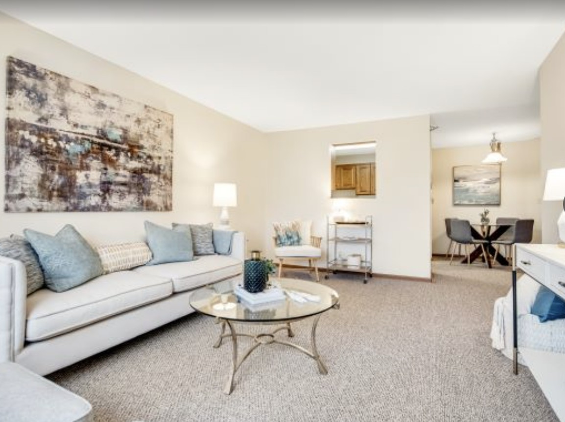 A cozy living room with neutral tones, featuring a sofa and glass coffee table, accented by artwork and soft lighting.