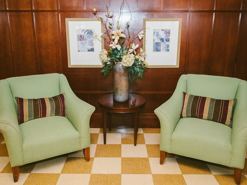 A cozy seating area featuring two green chairs with striped cushions, a wooden side table with a floral arrangement, and framed artwork on the wooden paneled wall.