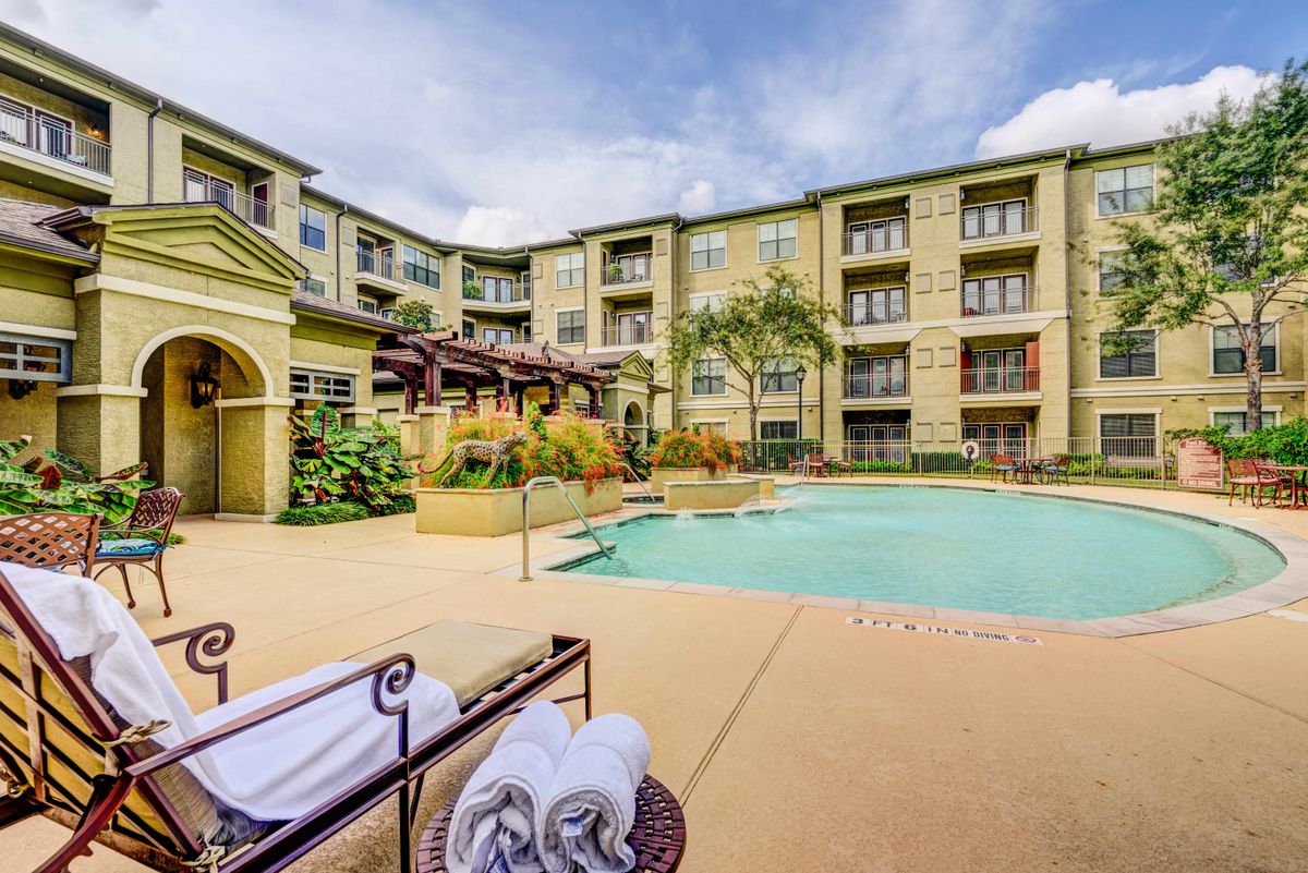 A modern apartment complex with a swimming pool surrounded by sun loungers and neatly folded towels, set on a sunny day.