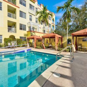A vibrant apartment complex features a swimming pool surrounded by cabanas and palm trees on a sunny day.