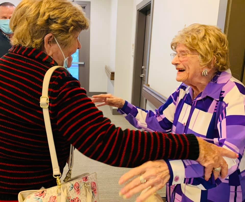 Two elderly women joyfully greet each other with open arms indoors.