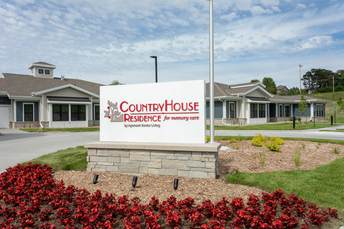 A sign for CountryHouse Residence surrounded by flowers with the building in the background.