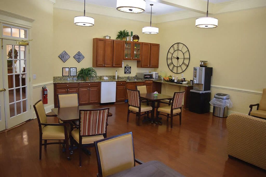 A cozy kitchen and dining area with wooden cabinets, tables, and chairs, featuring a large wall clock and decorative elements.