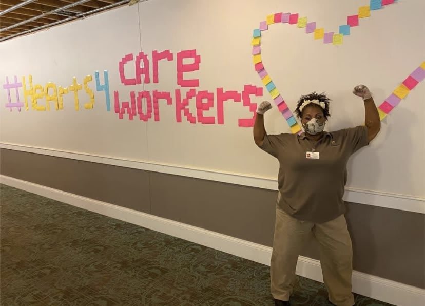 A healthcare worker poses in front of a wall adorned with post-it notes spelling 'care workers' and forming a heart shape.