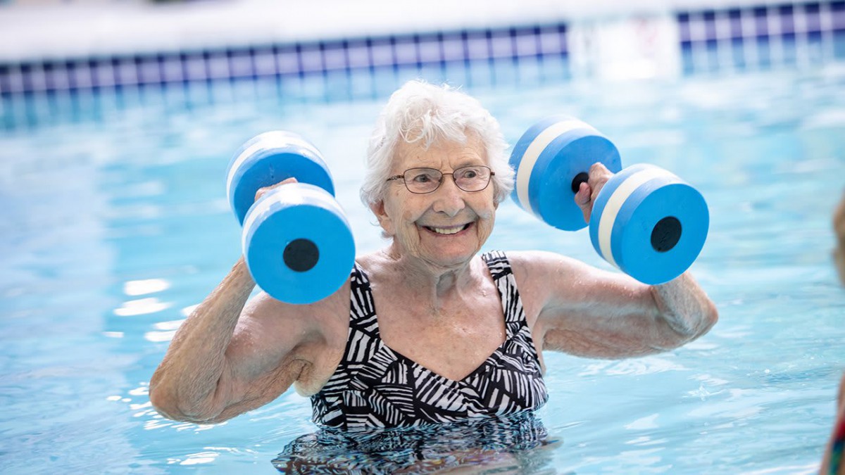 An elderly woman smiles while participating in a water exercise class using foam dumbbells in a pool.