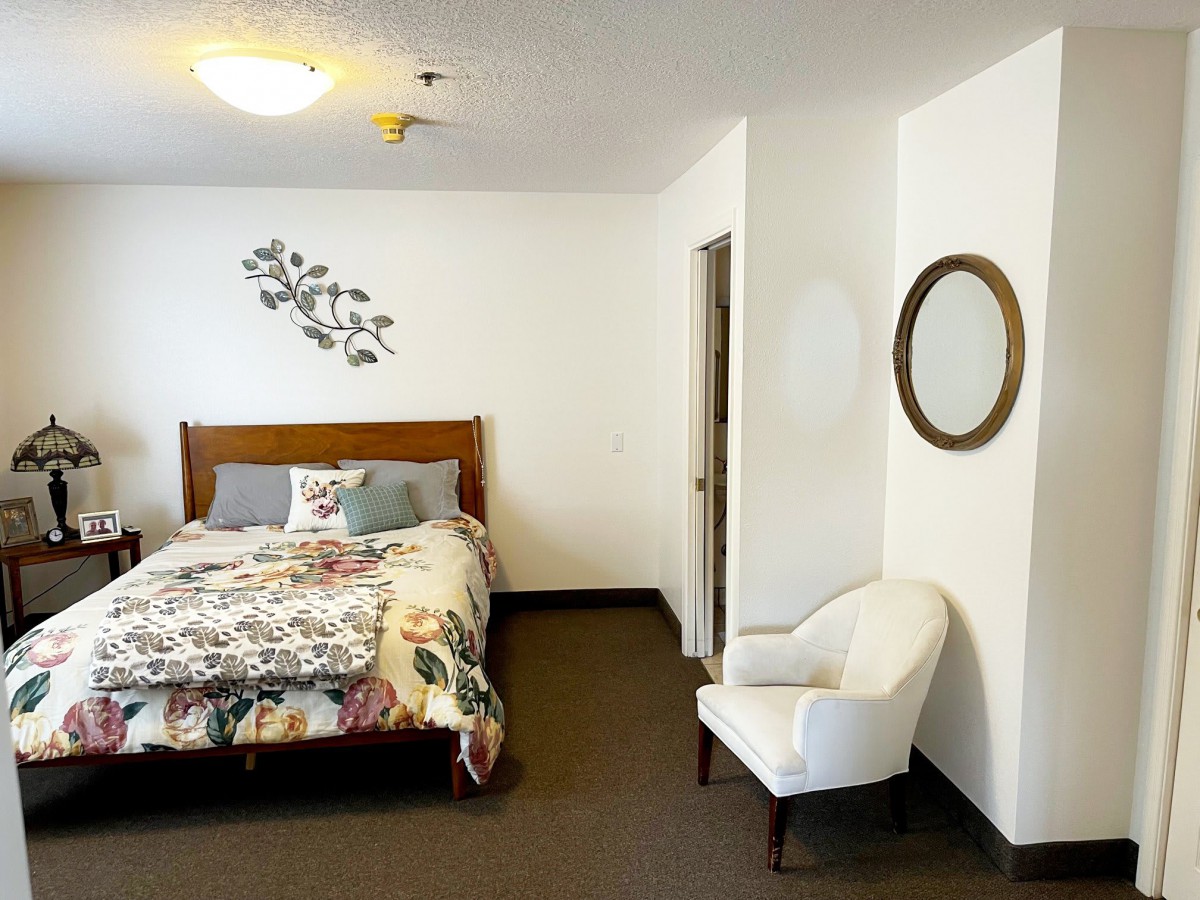 A cozy bedroom featuring a bed with a patterned comforter, an upholstered chair, and a wall-mounted mirror.