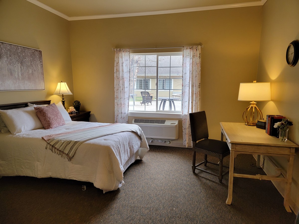 A cozy bedroom with a neatly made bed, bedside lamp, and a window letting in natural light.
