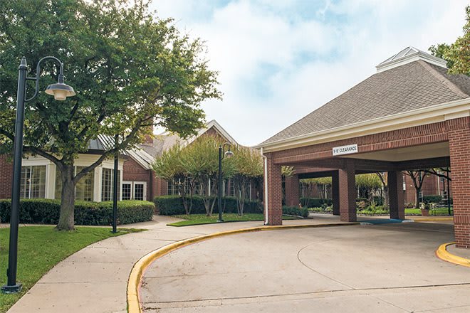 A building entrance with a driveway surrounded by trees and landscaping under a cloudy sky.