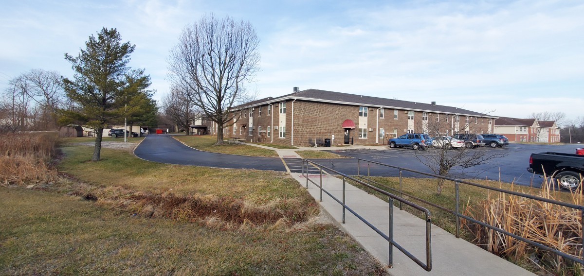 A brick building with a parking lot and barren trees, featuring a sidewalk and ramp in the foreground.