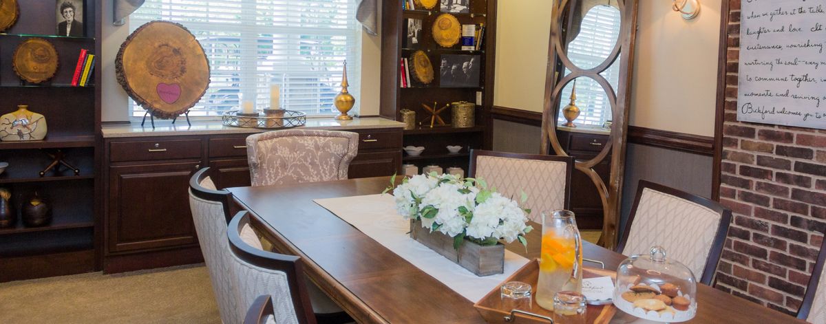 A cozy dining room with a wooden table, adorned with a floral centerpiece and decorative elements on surrounding shelves.