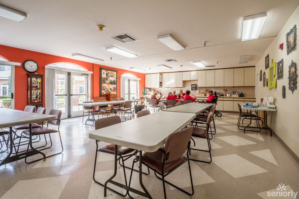 A spacious community center dining area with folding tables and chairs, featuring a kitchen space in the background.