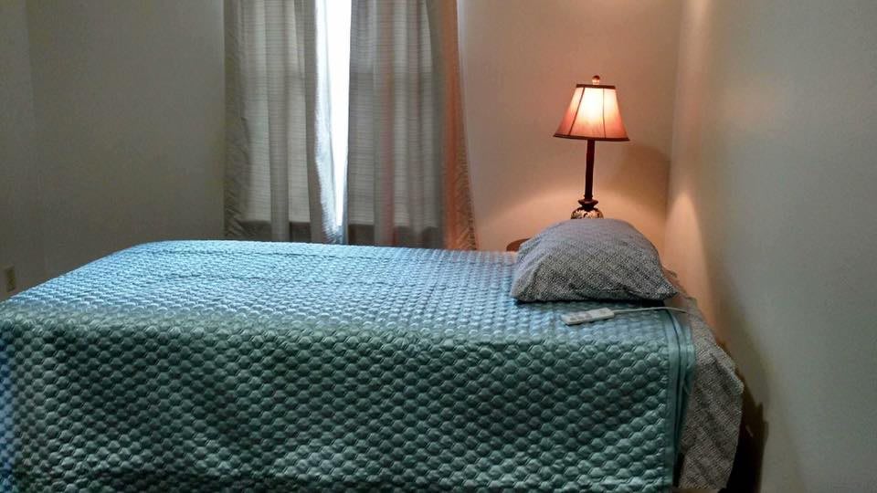 A neatly made bed with a textured blue coverlet beside a lit lamp.