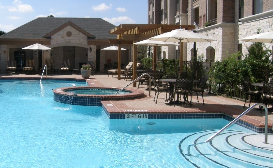 An outdoor swimming pool area with a jacuzzi, surrounded by patio furniture and umbrellas on a sunny day.