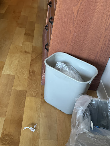 A gray trash can sits on a wooden floor, with some waste visible inside.