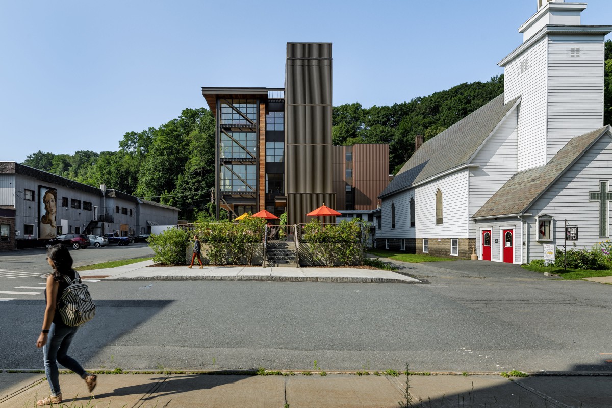 A view of a modern building with large glass windows adjacent to a traditional church, surrounded by greenery and outdoor seating under umbrellas.