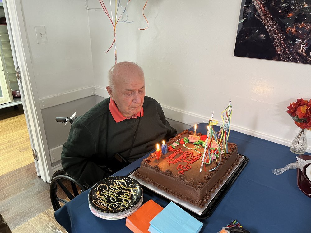 An elderly person sits in front of a birthday cake with lit candles, ready to celebrate.