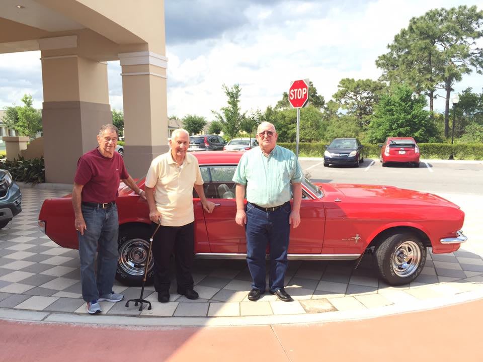 Three men standing in front of a vintage red car outside.