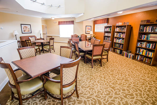 A cozy library with multiple tables and chairs, surrounded by bookshelves in a well-lit community room.