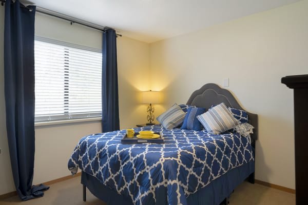 A cozy bedroom with a neatly made bed, featuring blue patterned bedding and pillows, a bedside lamp, and a window with blinds.