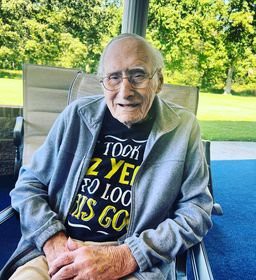 An elderly man sitting outdoors, wearing glasses and a t-shirt with a cheerful message.