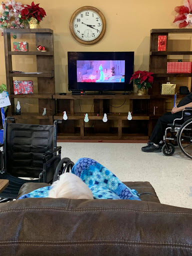 An elderly person wrapped in a blanket watching television in a care facility, with another individual in a wheelchair nearby.