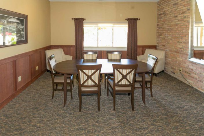 A dining room with a round table and six chairs, featuring a carpeted floor and a window with curtains.