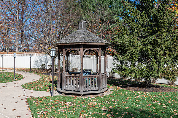 A wooden gazebo surrounded by trees and fallen autumn leaves in a garden setting.