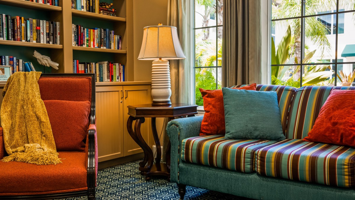 A cozy living room with a striped sofa, decorative pillows, a lamp on a side table, and a bookshelf by the window.