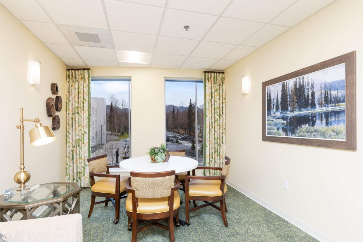 A cozy dining area with a round table surrounded by chairs, adorned with a plant and flanked by large windows and artwork.