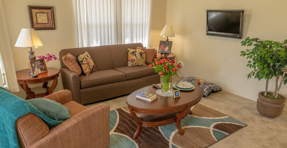 A cozy living room featuring a brown sofa, a wooden coffee table with flowers, and a television on the wall.