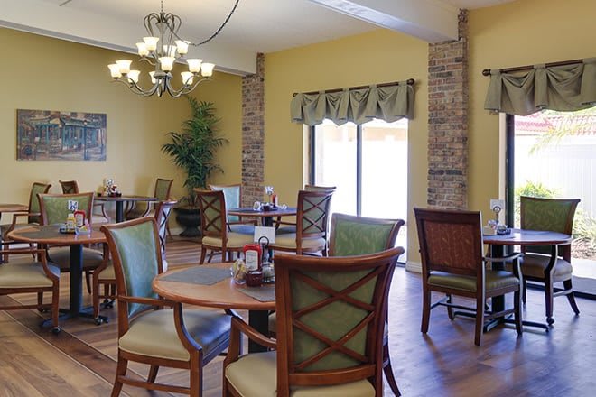 A cozy dining area with wooden tables and chairs, lit by a chandelier and natural light.