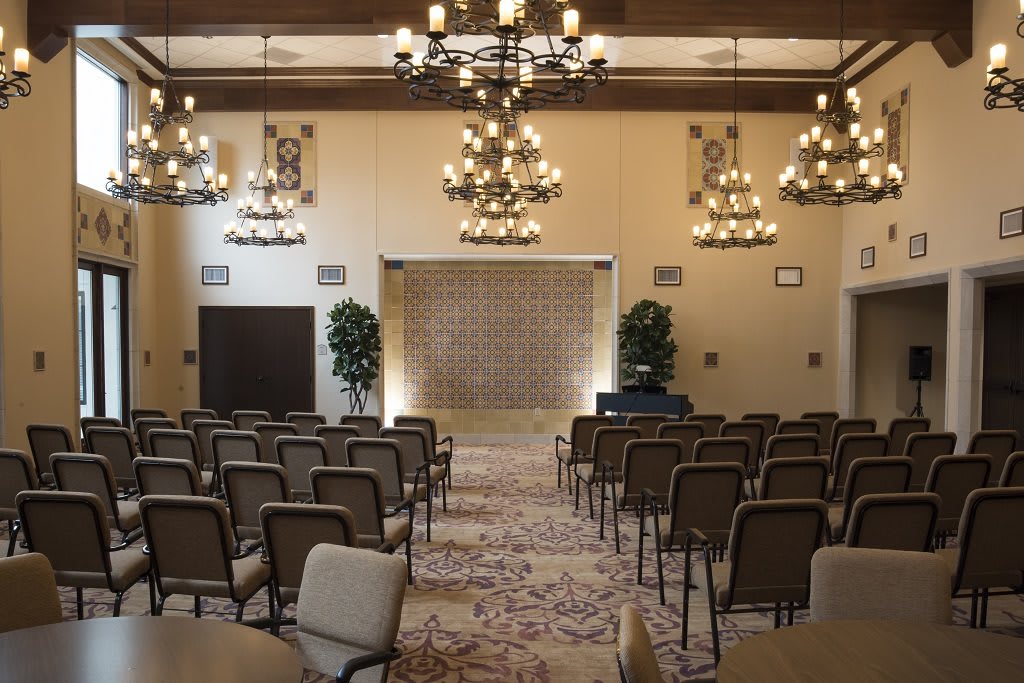 A well-lit conference room with rows of chairs facing a stage, adorned with chandeliers and decorative plants.