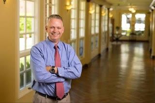 A man in business attire smiling in a well-lit office corridor.