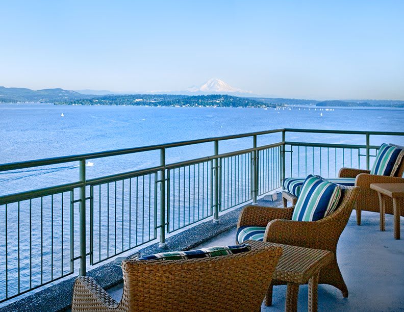 A balcony with comfortable seating offers a stunning view of the ocean and a distant mountain.