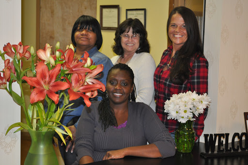 A group of four women posing together in an office setting, surrounded by beautiful flower arrangements.