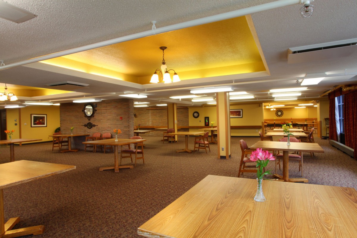 An empty dining hall with wooden tables and chairs, warm lighting, and decorative elements on the walls.