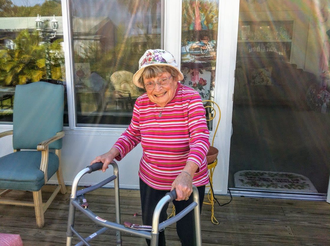 An elderly woman with a walker, wearing a striped shirt and hat, stands on a porch.